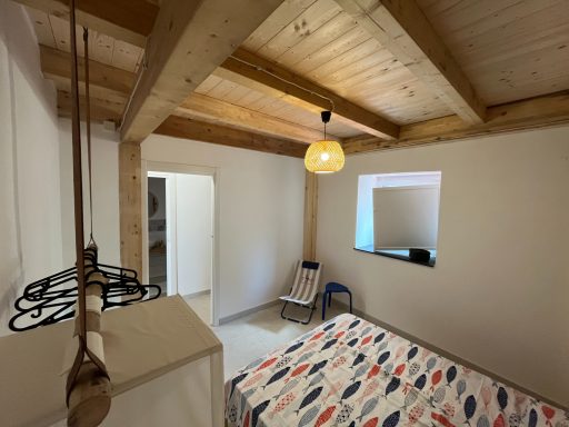 Helle Zimmer mit Holzdecken, einem Bett, einem Stuhl und Zugang zu einem angrenzenden Raum.